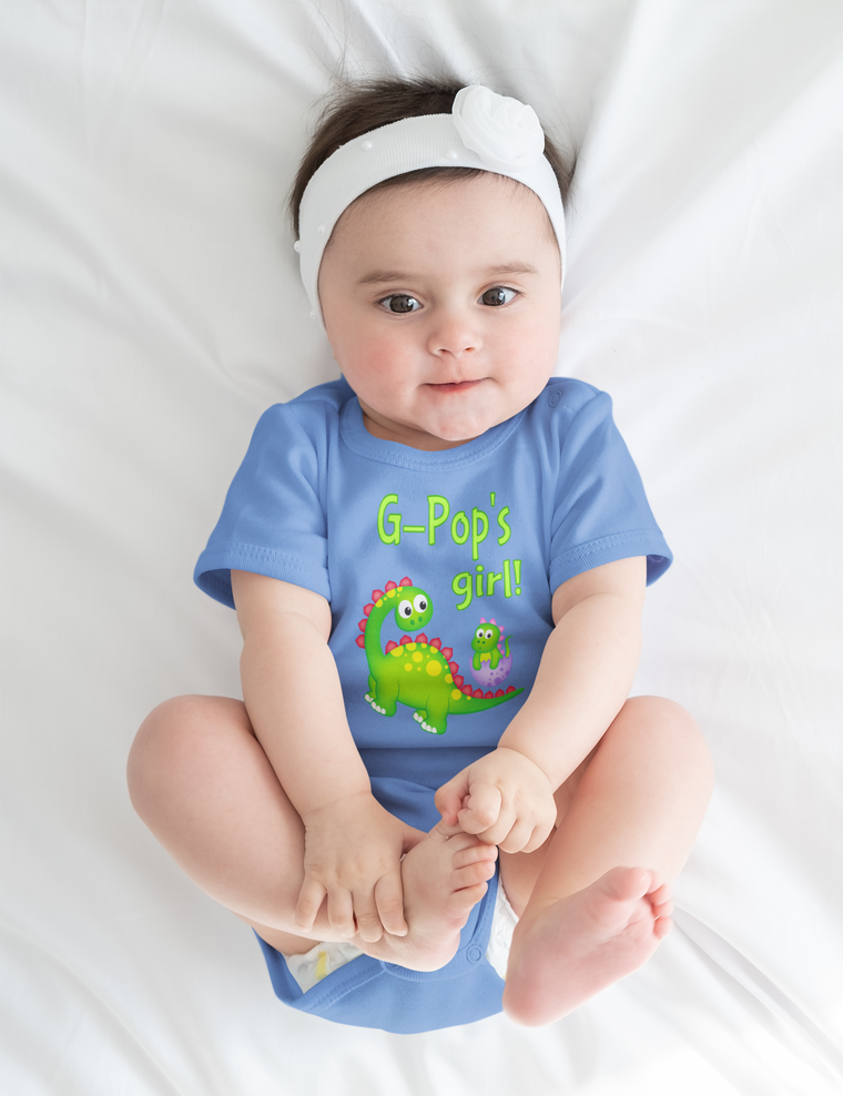 A baby girl wearing a blue onesie with a dinosaur design and the text "G-Pop's girl!" is lying on a white bed.