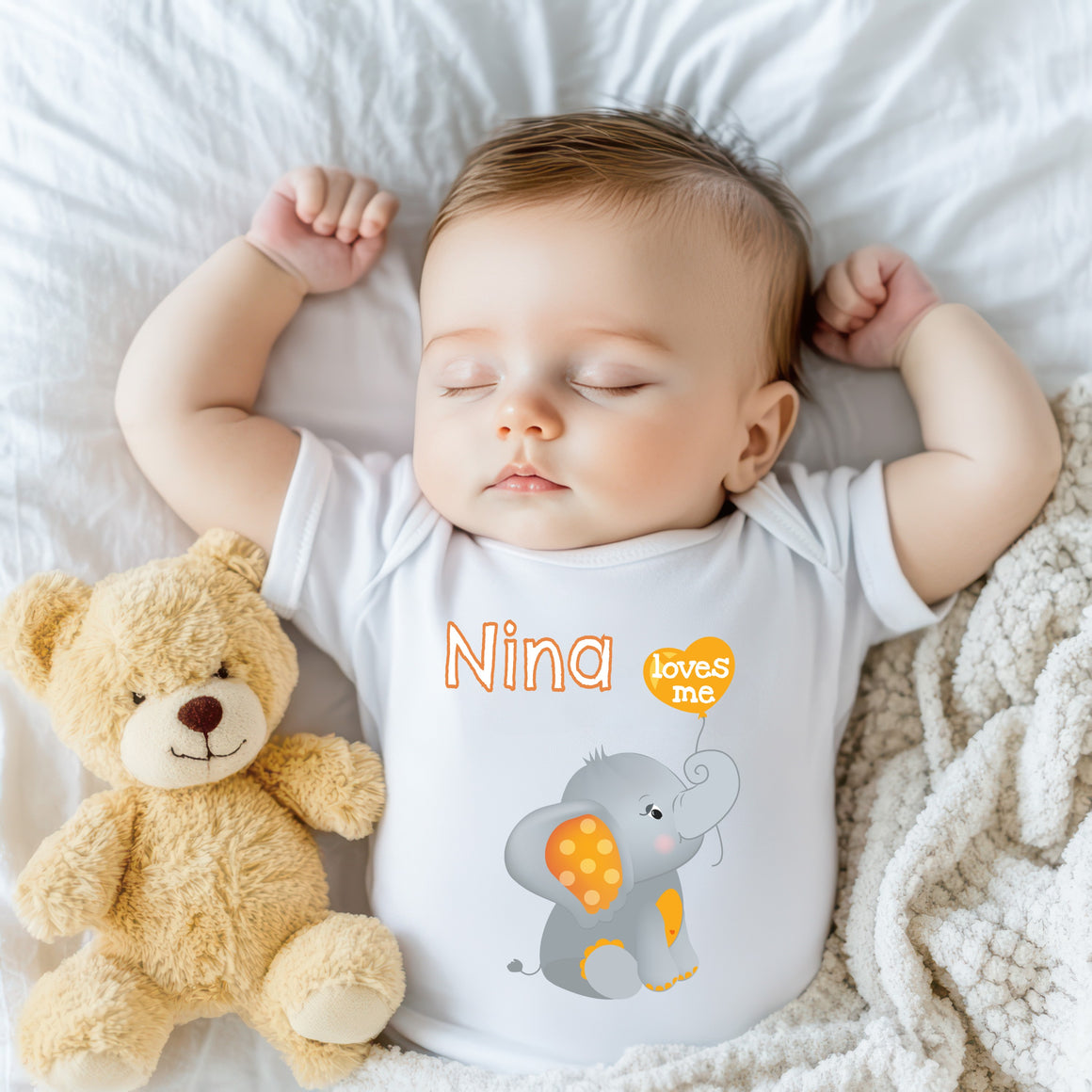 A sleeping baby wearing a white onesie with an elephant and the name "Nina" printed on it, accompanied by a teddy bear.