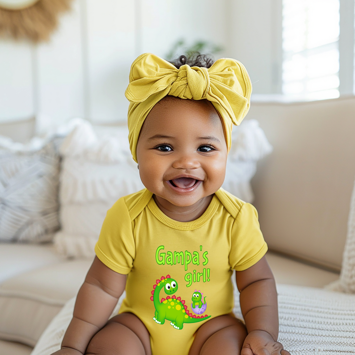 A smiling baby girl wearing a yellow onesie with a dinosaur design and a yellow headband.