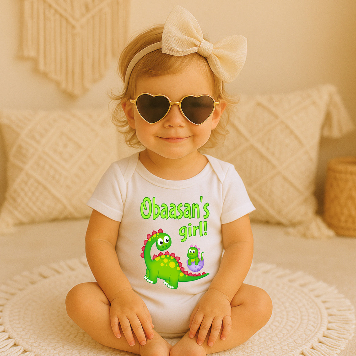 A young girl wearing a white shirt with a dinosaur graphic and heart-shaped sunglasses sits on a white rug.