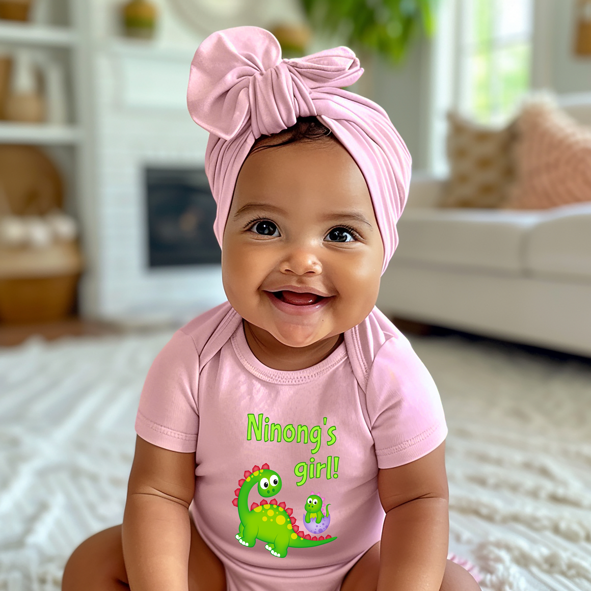 A smiling baby girl wearing a pink headband and a pink onesie with a dinosaur design, sitting on a white rug in a living room.