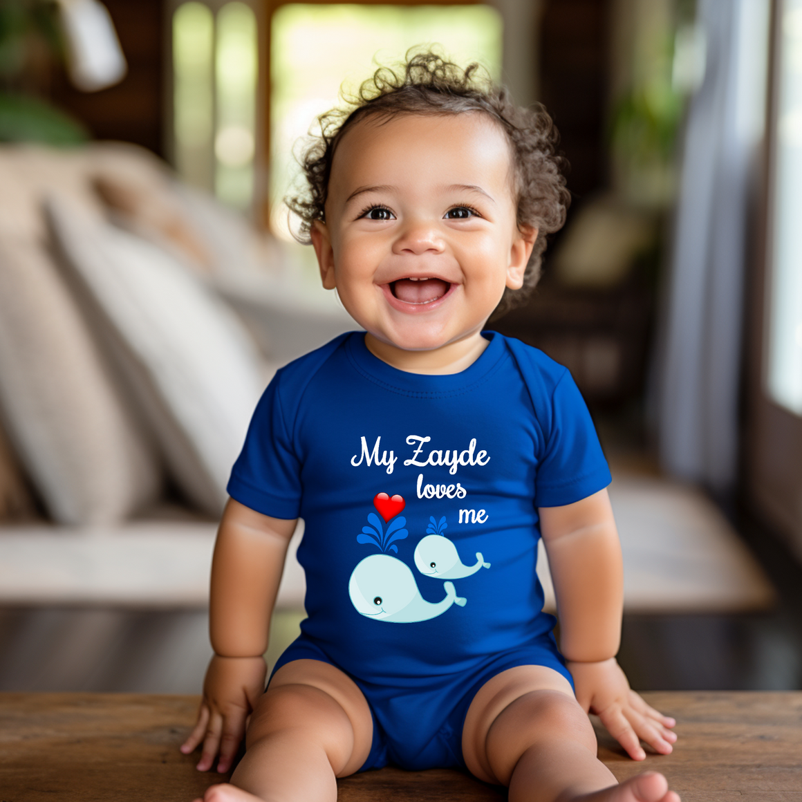A smiling baby boy wearing a blue onesie with a whale design and the text "My Zayde loves me" is sitting on a wooden surface.