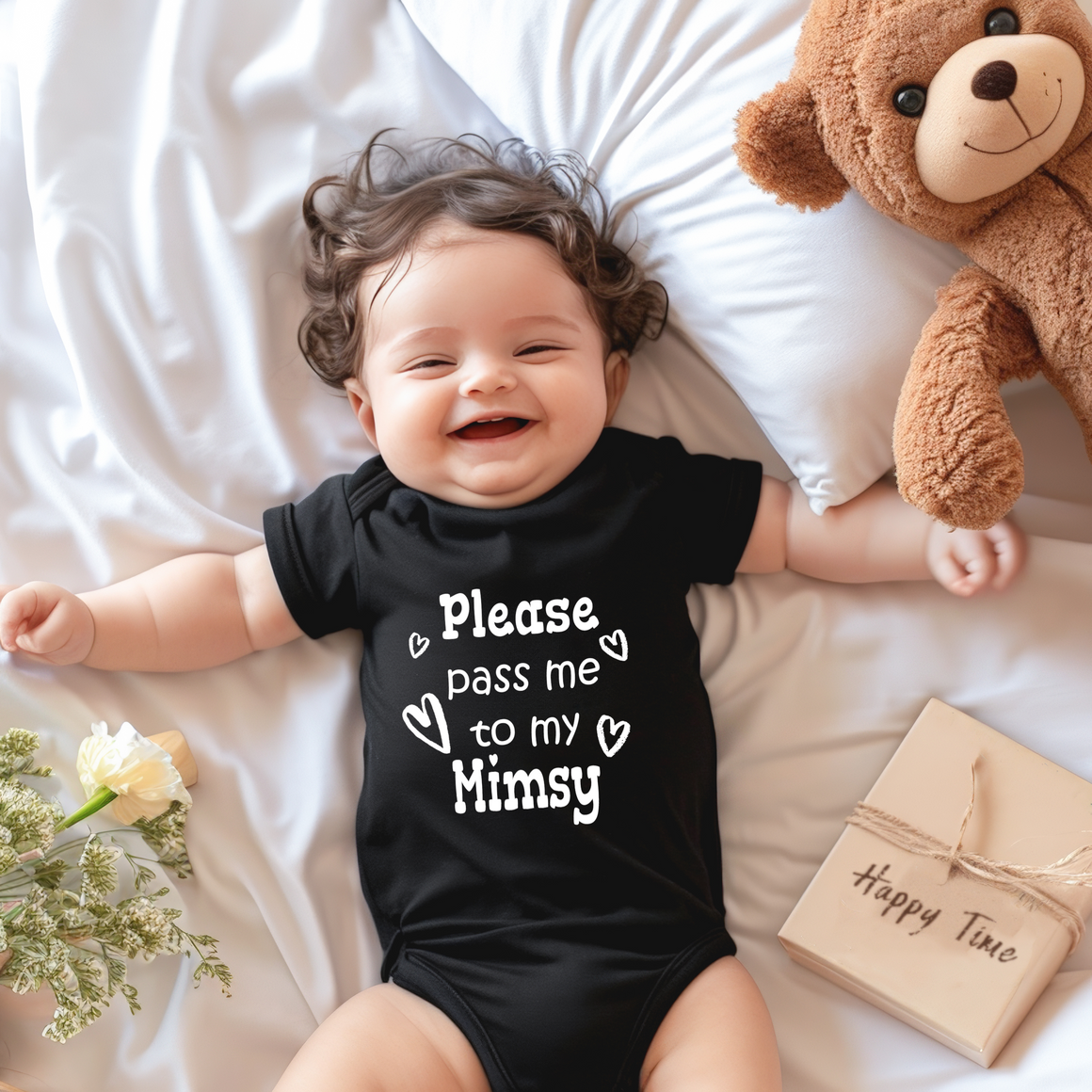 A smiling baby wearing a black onesie with the text "Please pass me to my Mimsy" lies on a bed next to a teddy bear and a gift box.