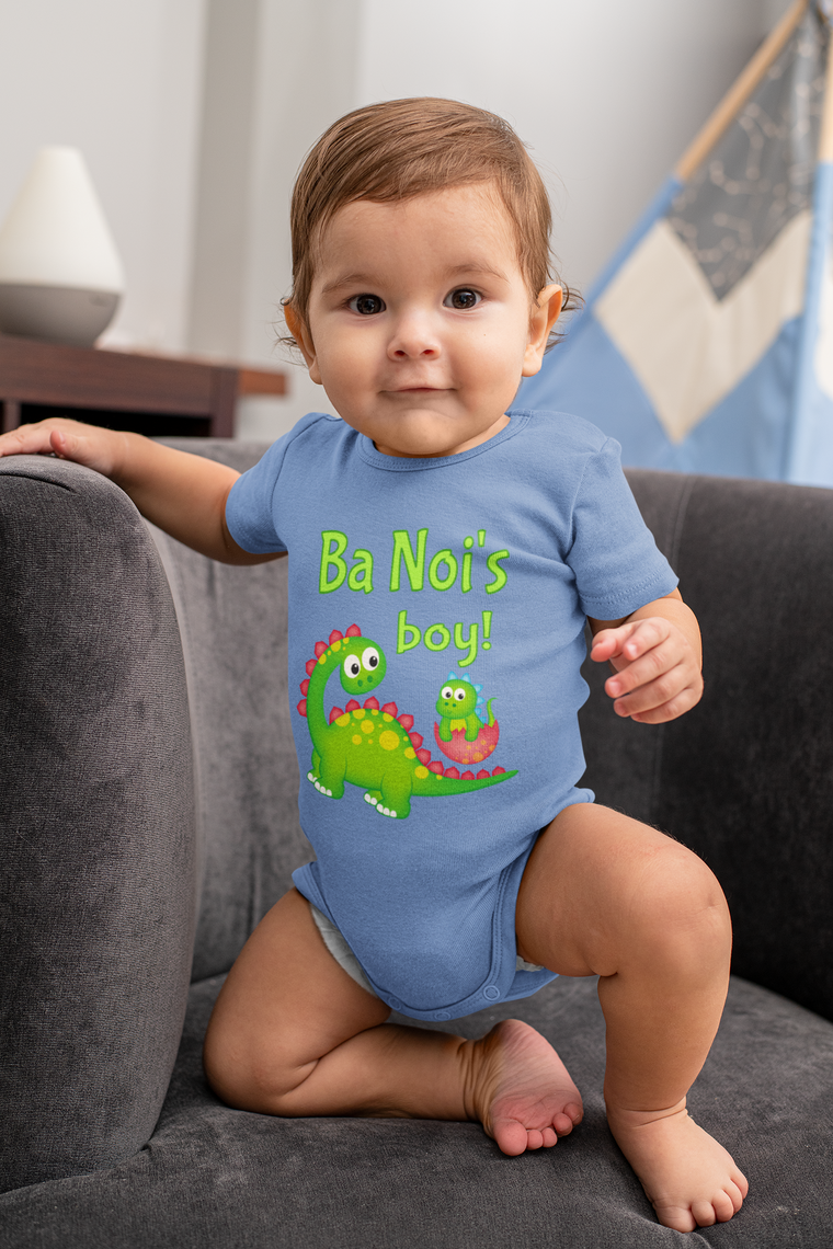 A smiling baby boy wearing a blue onesie with a cartoon dinosaur and the text "Ba Noi's boy!" is sitting on a couch.