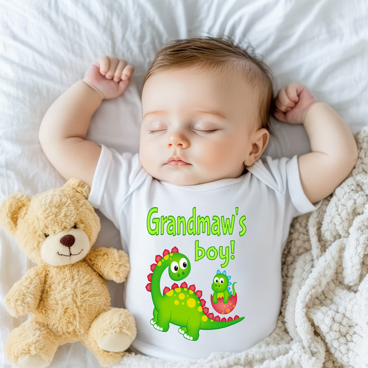 A sleeping baby wearing a white onesie with a dinosaur graphic and the text "Grandma's boy!" next to a teddy bear.