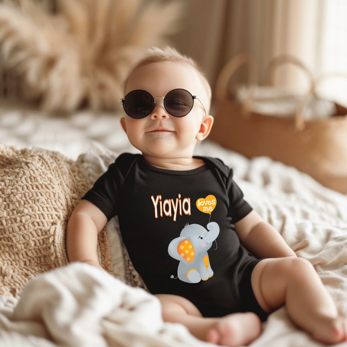 A baby wearing a black shirt with a cartoon elephant and the word "Yiayia" printed on it, sitting on a bed and wearing sunglasses.