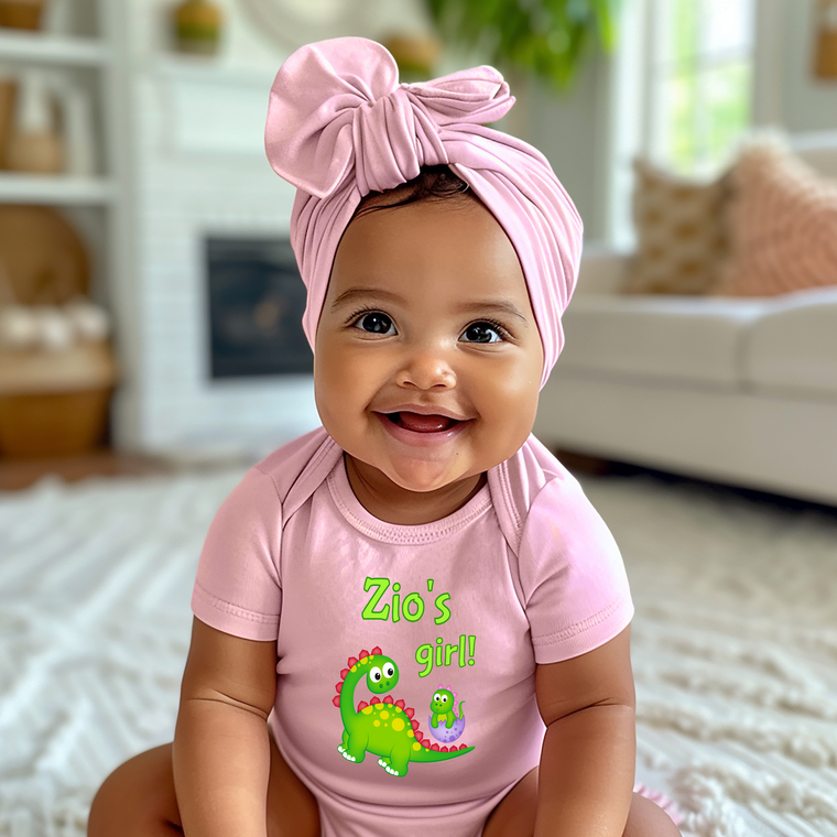 A smiling baby girl wearing a pink headband and a pink onesie with a dinosaur design, sitting on a white rug in a living room.