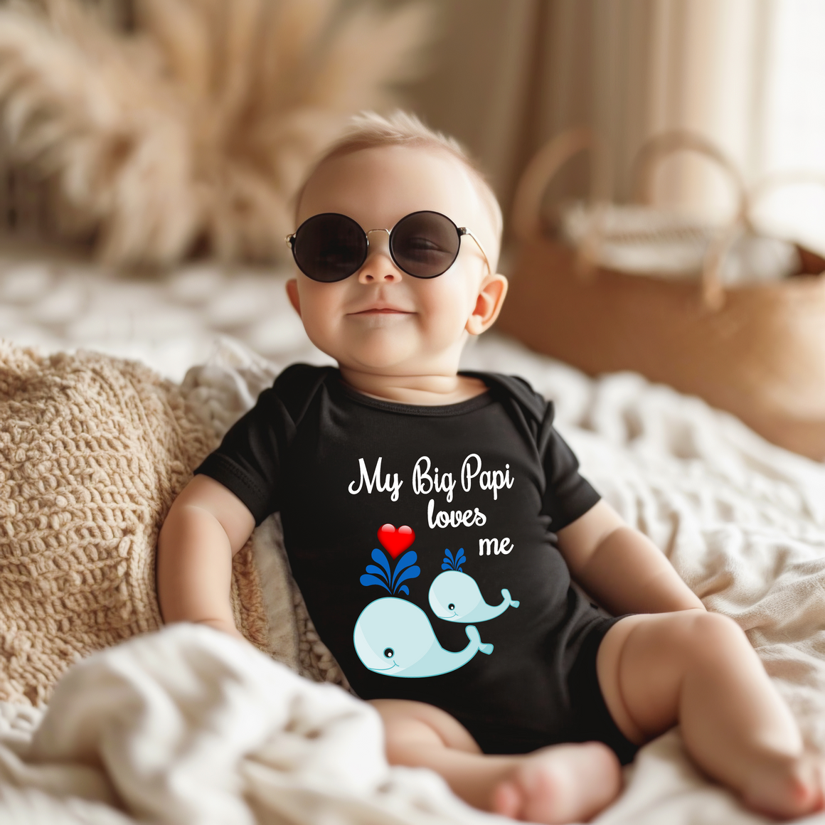 A baby wearing a black shirt with a graphic of a whale and the text "My Big Papi loves me" is sitting on a bed.