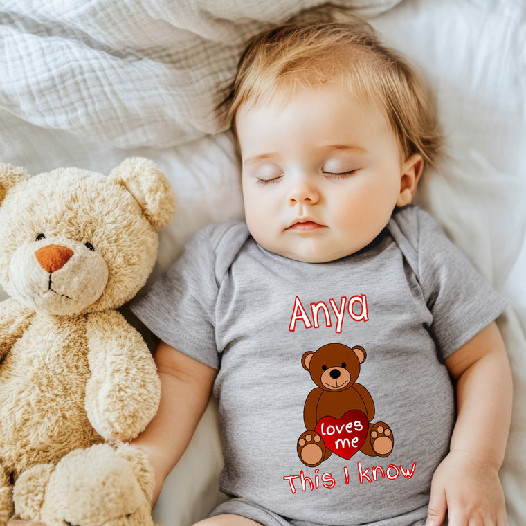 A baby sleeping next to a teddy bear, wearing a gray onesie with a personalized name and a heart-shaped design.