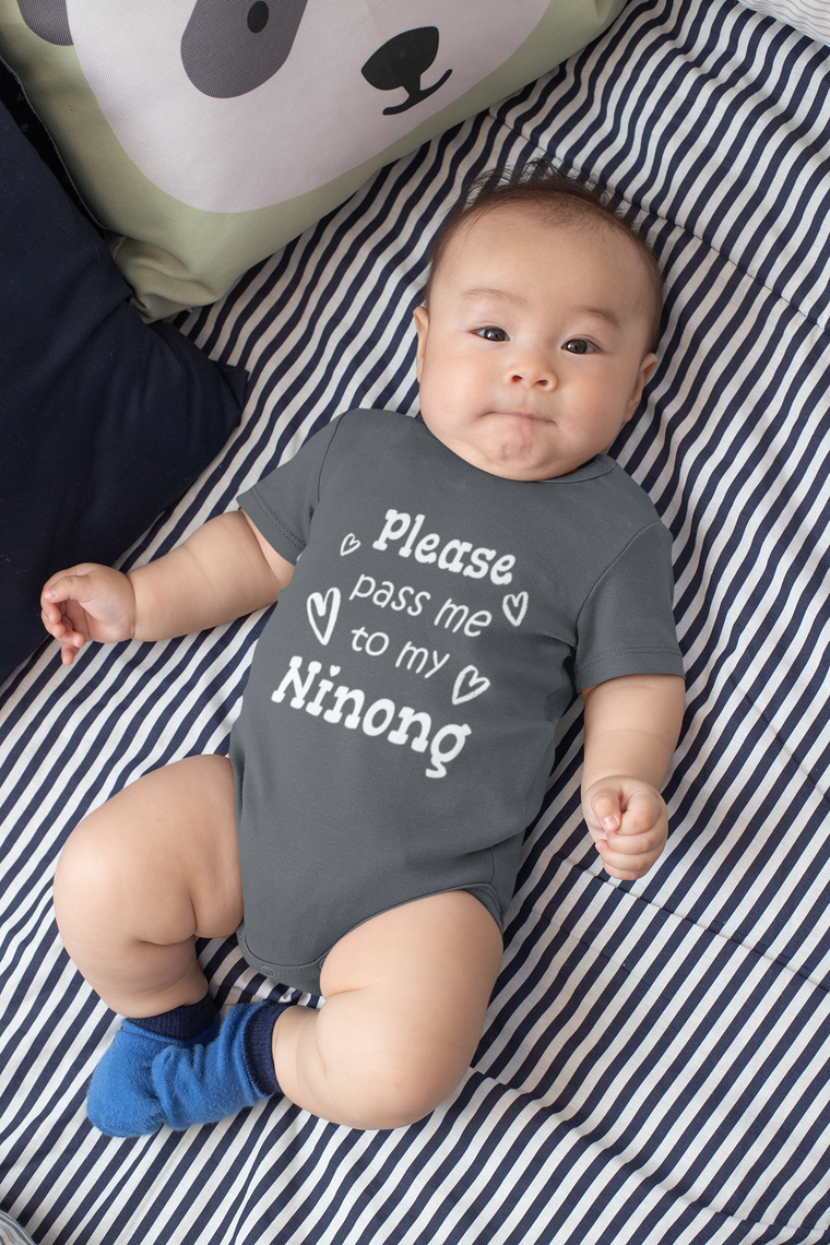 A baby wearing a grey onesie with the text "Please pass me to my Ninong" lying on a striped bed.