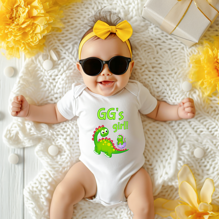 A baby wearing a white onesie with a dinosaur and the text "GG's girl!" lying on a white blanket surrounded by yellow flowers.