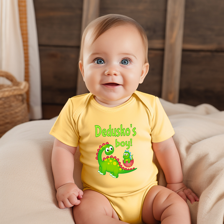 A smiling baby wearing a yellow onesie with a dinosaur graphic and the text "Dedushka's boy!"