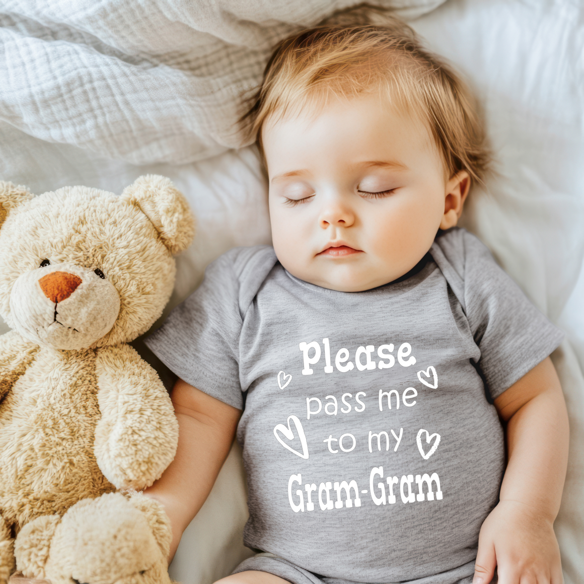 A sleeping baby wearing a gray onesie with the text "Please pass me to my Gram-Gram" is lying next to a teddy bear on a bed.