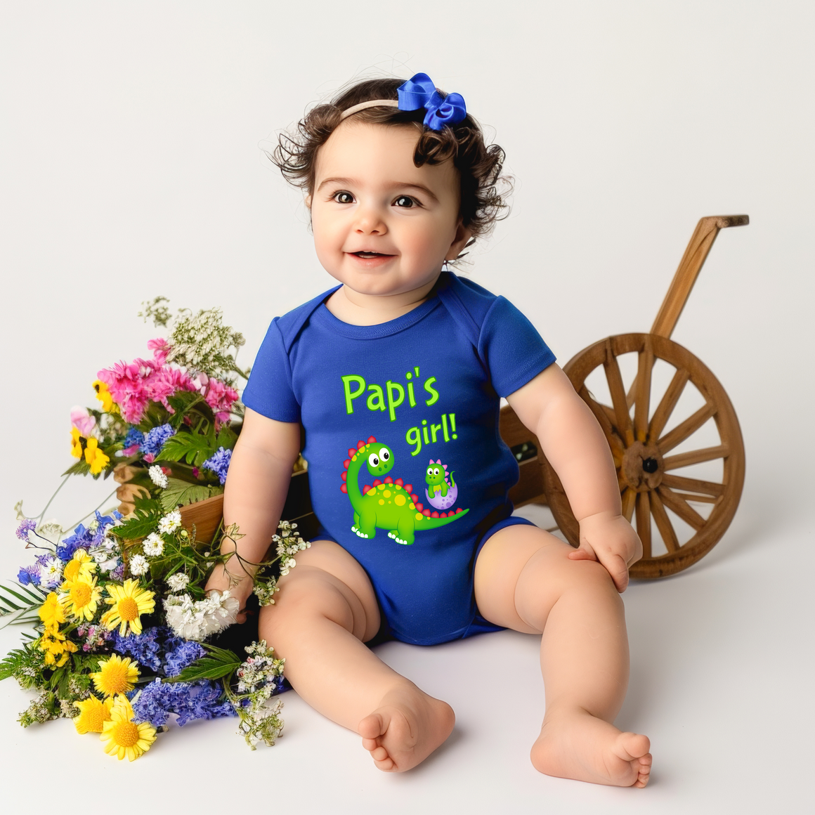 A smiling baby girl wearing a blue onesie with a dinosaur and the text "Papi's girl!" is sitting next to a wooden wagon wheel and a basket filled with colorful flowers.
