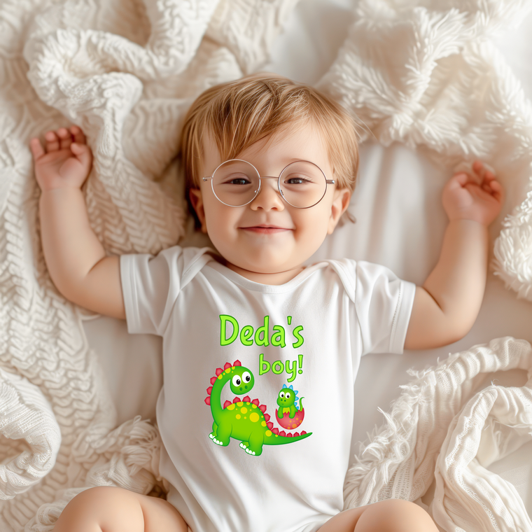 A smiling baby wearing a white onesie with a dinosaur graphic and the text "Dada's boy!" lying on a white blanket.