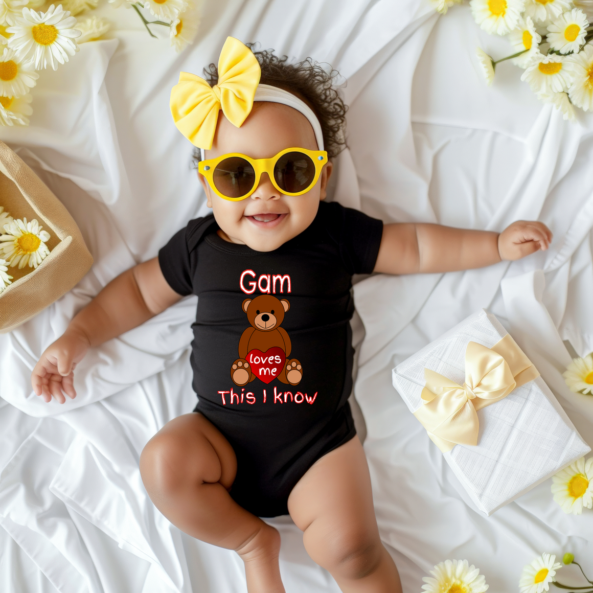 A smiling baby girl wearing a black outfit with a teddy bear and sunglasses lying on a bed surrounded by yellow flowers.