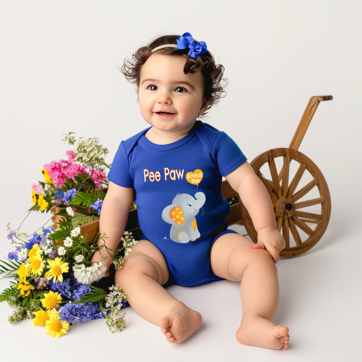 A smiling baby girl wearing a blue onesie with an elephant design sits next to a wooden wagon filled with colorful flowers.