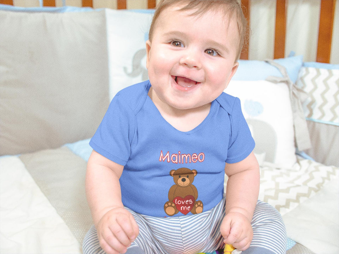 A smiling baby boy wearing a blue shirt with a teddy bear graphic and the word "Maimeo" printed on it, sitting on a bed.