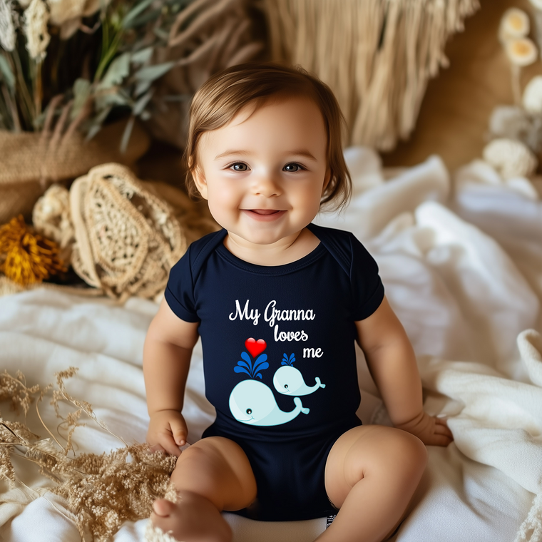 A smiling baby wearing a navy blue onesie with a whale design and the text "My Grandma loves me" is sitting on a bed with a floral background.