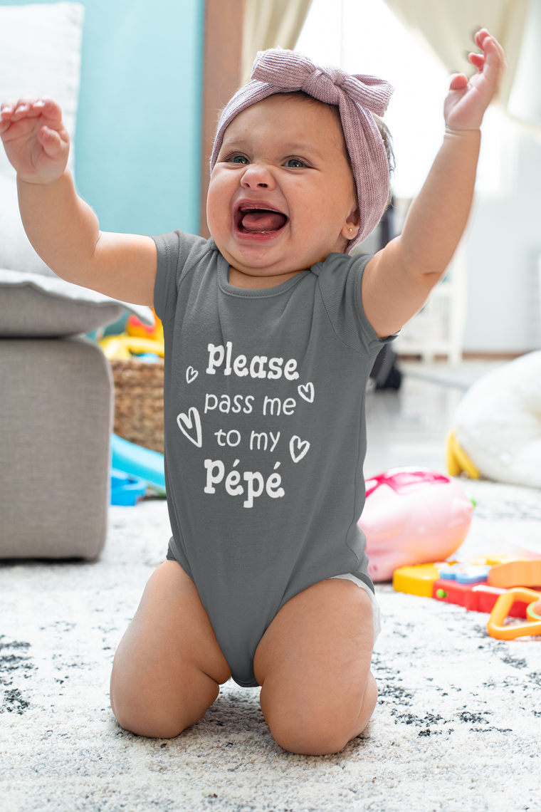 A smiling baby girl wearing a grey onesie with the text "Please pass me to my Pepe" is sitting on the floor with her arms raised in a joyful expression.