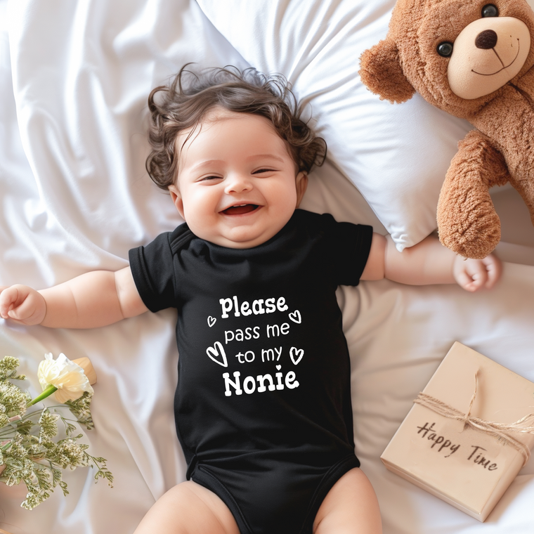 A smiling baby wearing a black onesie with text on it lies on a bed next to a teddy bear and a gift box.