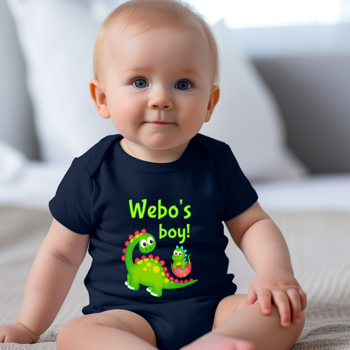 A smiling baby wearing a black t-shirt with a dinosaur graphic and text that says "Web's boy!".