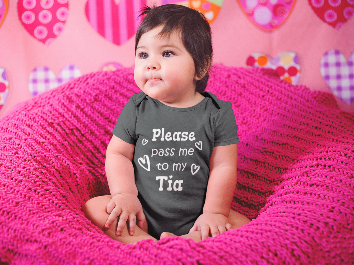 A baby wearing a gray t-shirt with the text "Please pass me to my Tia" sitting on a pink blanket.