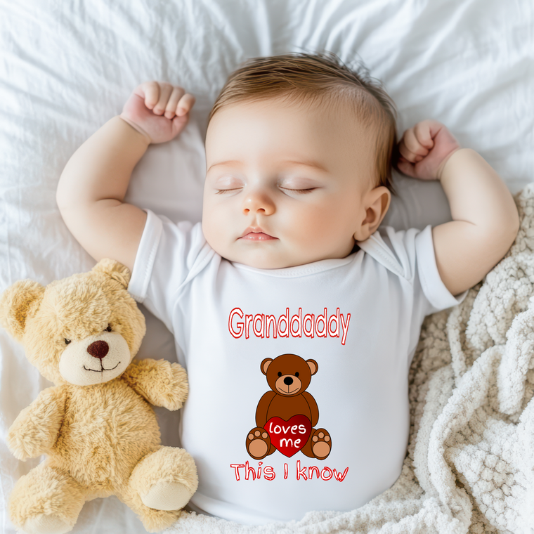 A sleeping baby wearing a white onesie with a teddy bear next to them.