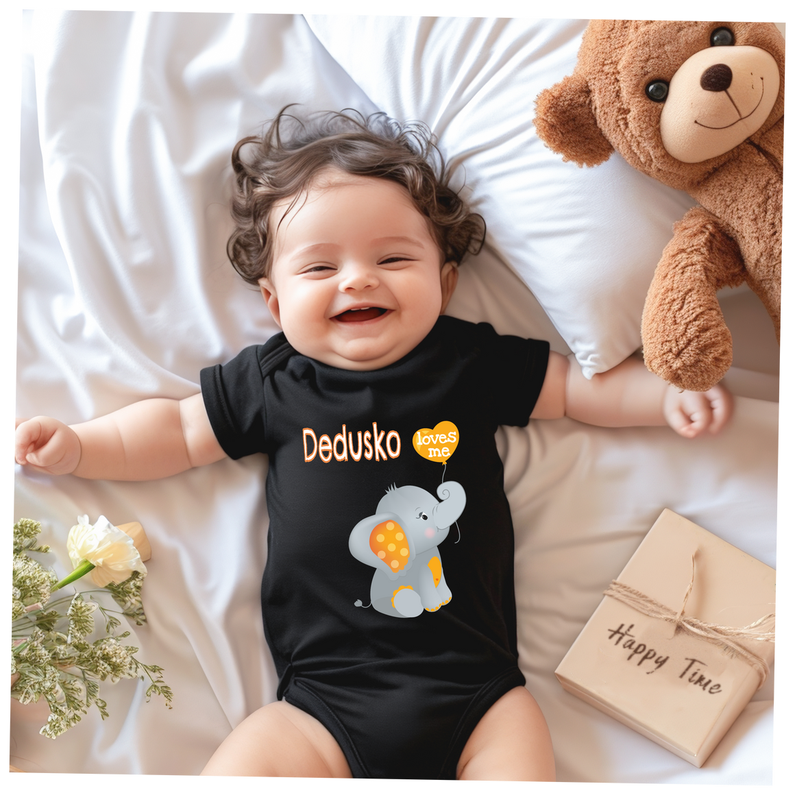 A smiling baby wearing a black t-shirt with an elephant design lies on a bed next to a teddy bear and a bouquet of flowers.