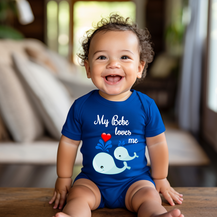 A smiling baby boy wearing a blue shirt with a whale design and the text "My Bebe loves me" is sitting on a wooden surface.
