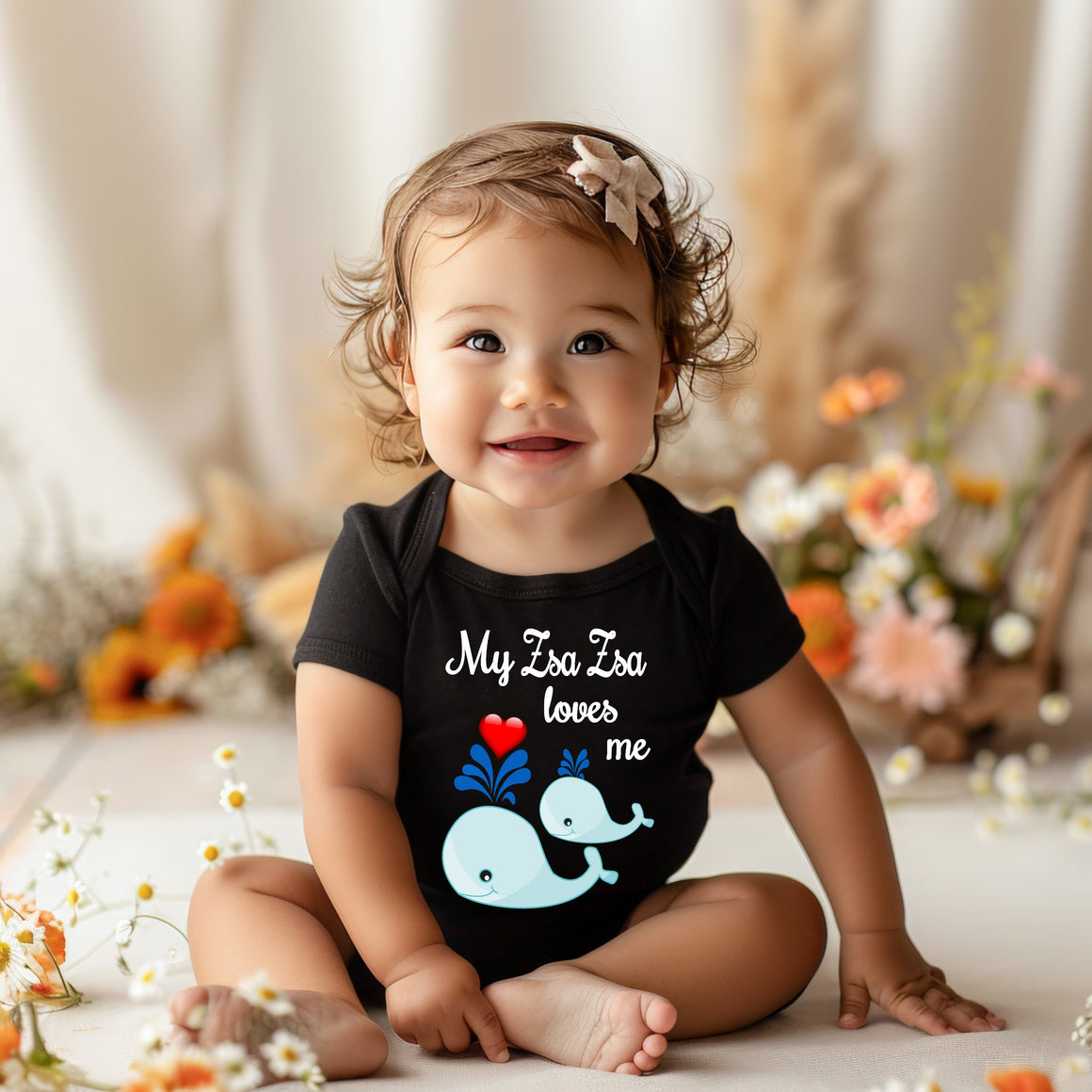 A smiling baby girl wearing a black shirt with a whale design and the text "My Za Za loves me" is sitting on the floor surrounded by flowers.