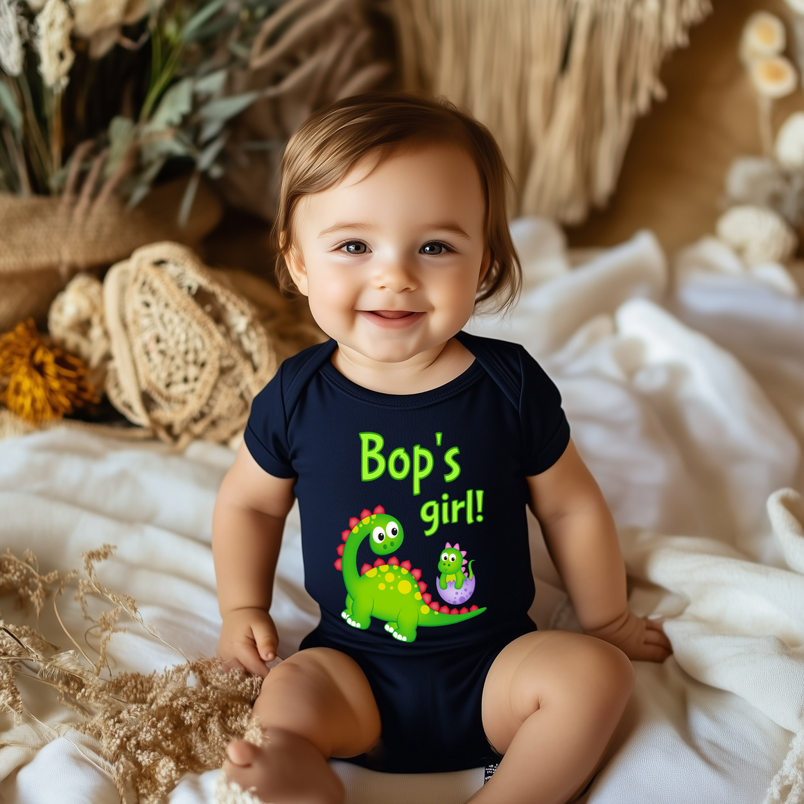 A smiling baby wearing a black t-shirt with a dinosaur graphic and the text "Bop's girl!"