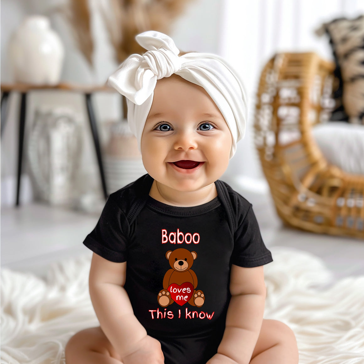 A smiling baby girl wearing a black t-shirt with a teddy bear graphic and text, sitting on a white blanket.
