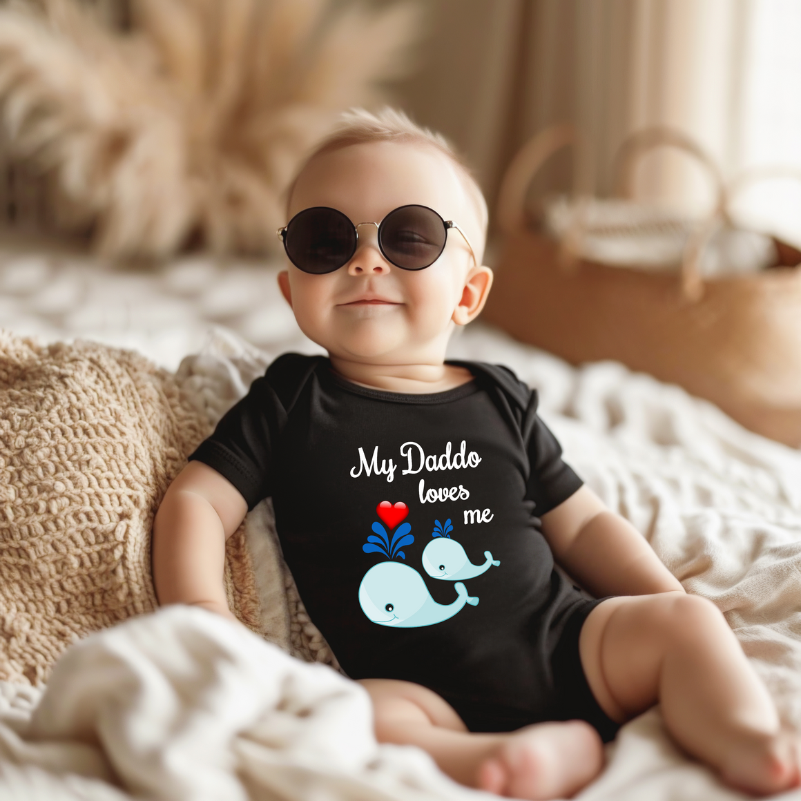 A baby wearing a black onesie with a graphic of a whale and the text "My Daddo loves me" is sitting on a bed.