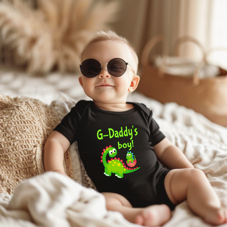A baby wearing a black onesie with a dinosaur graphic and the text "G-Daddy's boy!" is sitting on a bed.