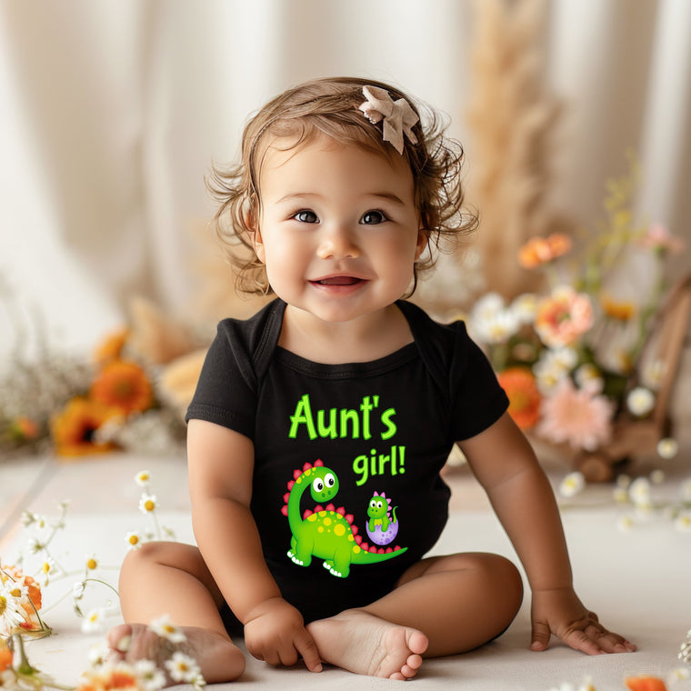 A smiling baby girl wearing a black shirt with a dinosaur and the text "Aunt's girl!" is sitting on the floor surrounded by flowers.