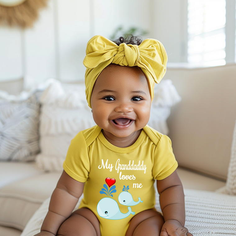 A smiling baby girl wearing a yellow outfit with a bow and a t-shirt that says "My Grandaddy loves me" sits on a couch.