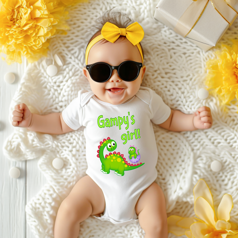 A baby wearing a white onesie with a dinosaur design and sunglasses lying on a white blanket surrounded by yellow flowers.