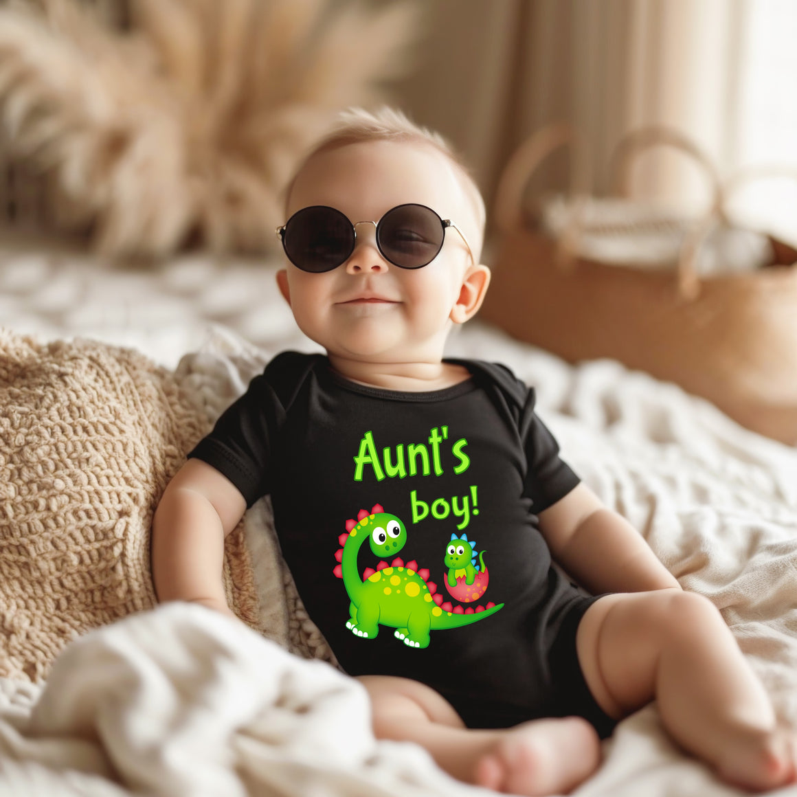 A baby wearing a black shirt with a dinosaur graphic and the text "Aunt's boy!" is sitting on a bed, wearing sunglasses.