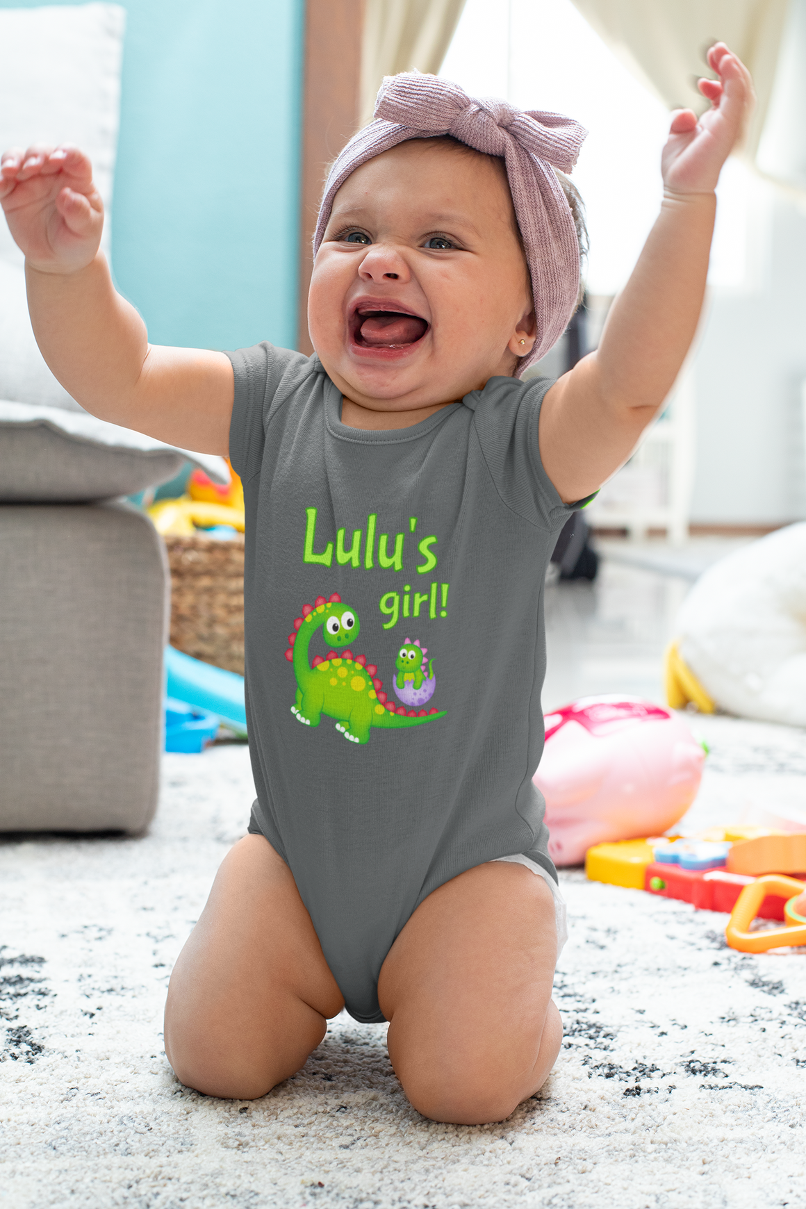 A smiling baby girl wearing a gray onesie with a dinosaur design and the text "Lulu's girl!" is sitting on the floor with her arms raised in a joyful expression.