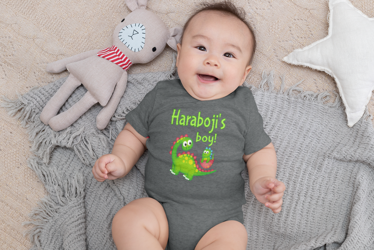 A baby wearing a gray onesie with a dinosaur design and the text "Haraboni's boy!" lying on a gray blanket next to a stuffed animal.