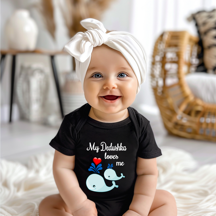 A smiling baby girl wearing a black t-shirt with a whale graphic and text, sitting on a white blanket.