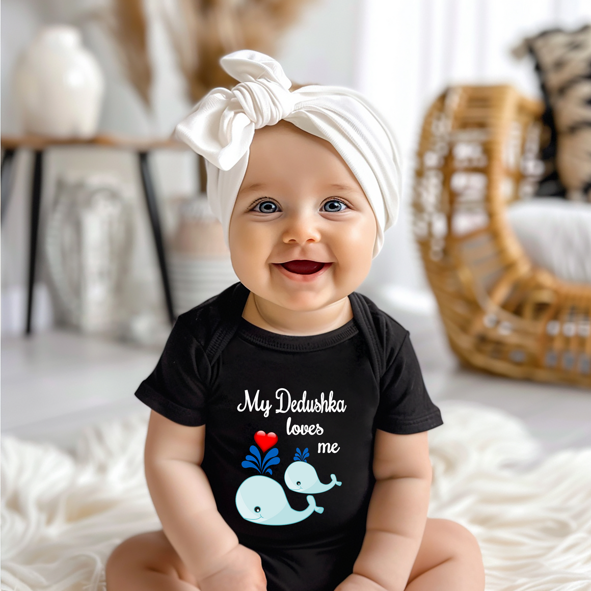 A smiling baby girl wearing a black t-shirt with a whale graphic and text, sitting on a white blanket.