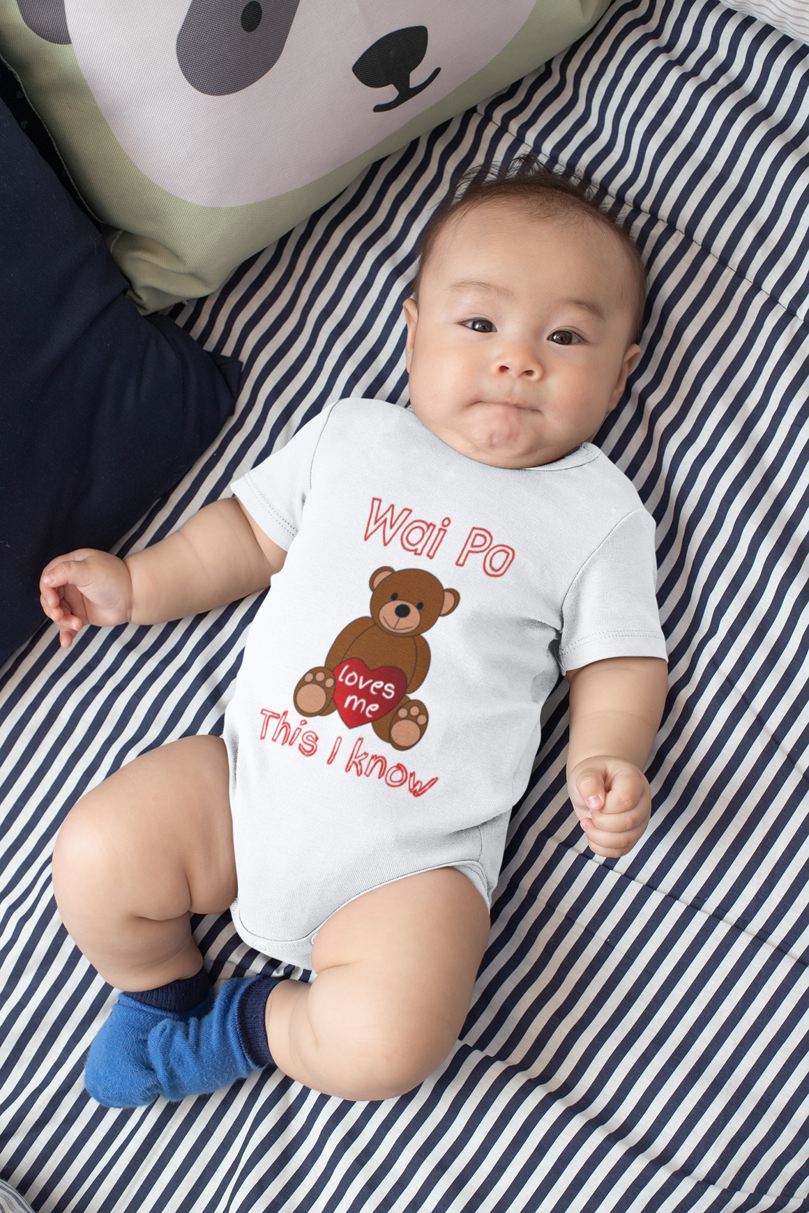 A baby wearing a white onesie with a teddy bear graphic and text, lying on a striped bed.