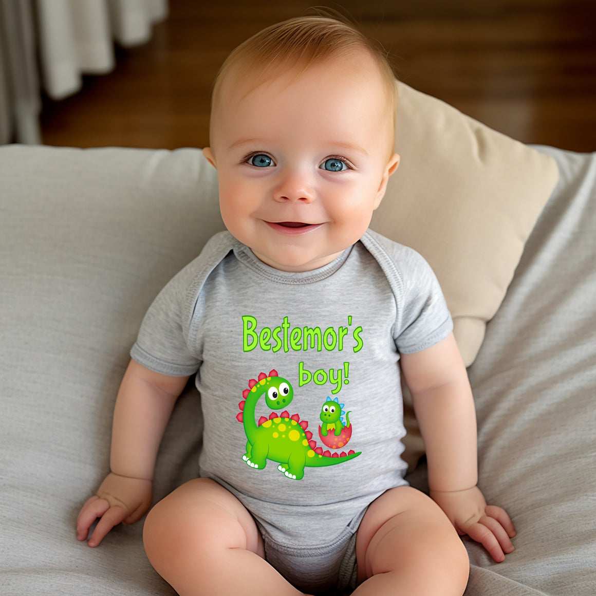 A smiling baby boy wearing a gray onesie with a green dinosaur and the text "Bestemor's boy!"