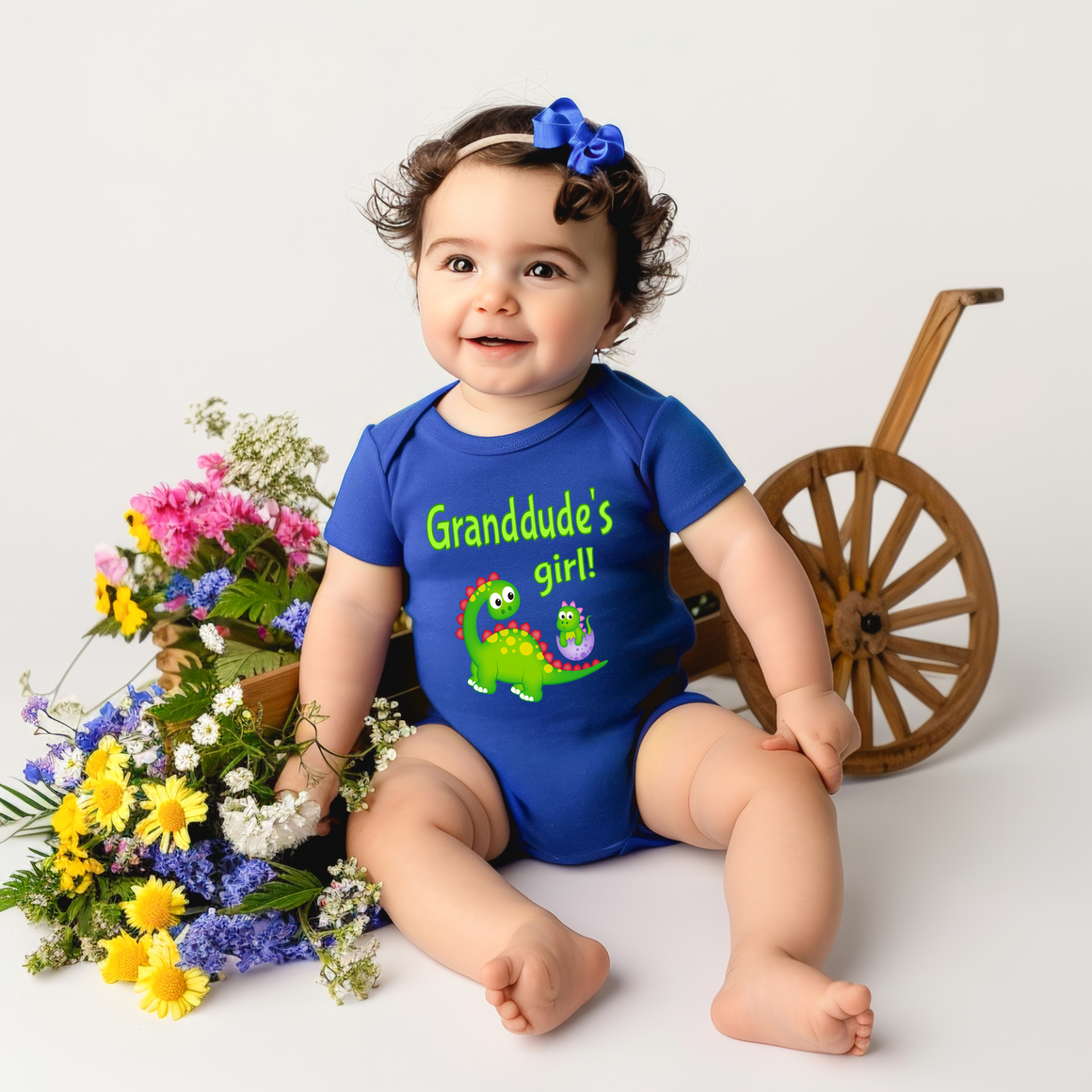 A smiling baby girl wearing a blue onesie with a dinosaur graphic sits next to a wooden toy wagon filled with colorful flowers.