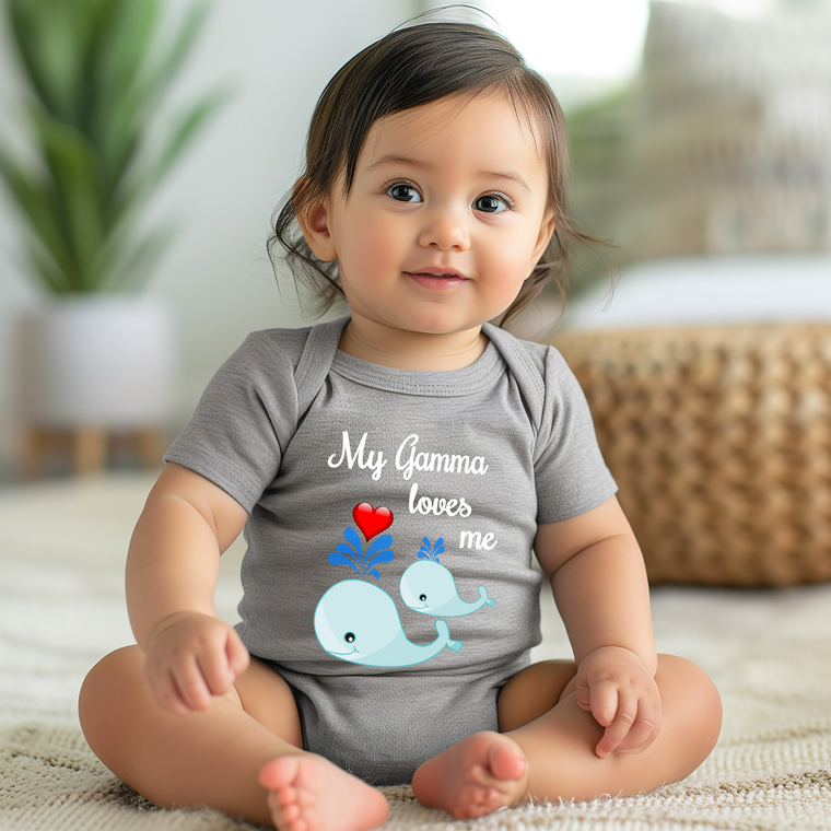A smiling baby girl wearing a gray onesie with a graphic of a whale and the text "My Gma loves me" is sitting on the floor.