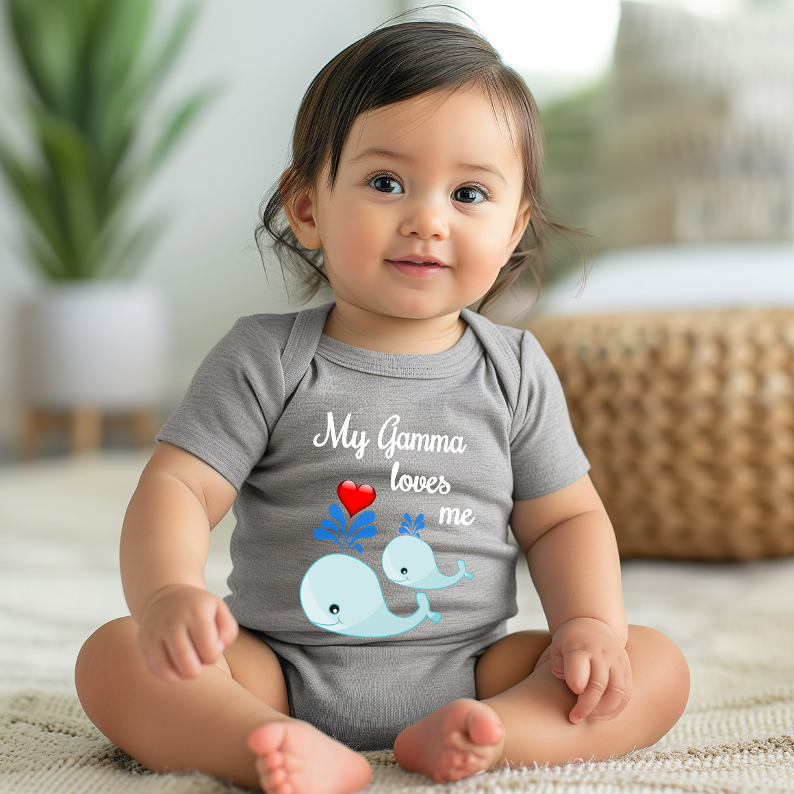A smiling baby girl wearing a gray onesie with a graphic of a whale and the text "My Gma loves me" is sitting on the floor.