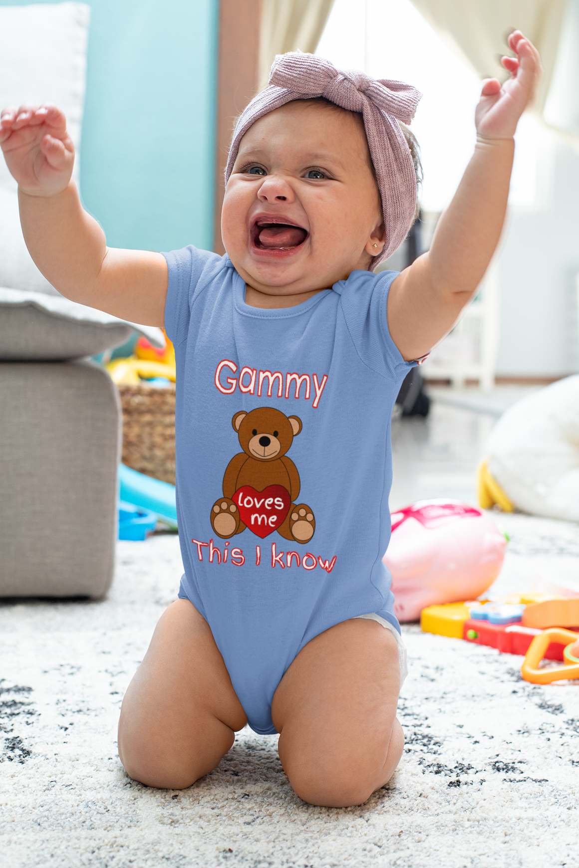 A smiling baby girl wearing a blue onesie with a teddy bear graphic and the text "Gamy loves me This I know" is sitting on the floor with her arms raised.