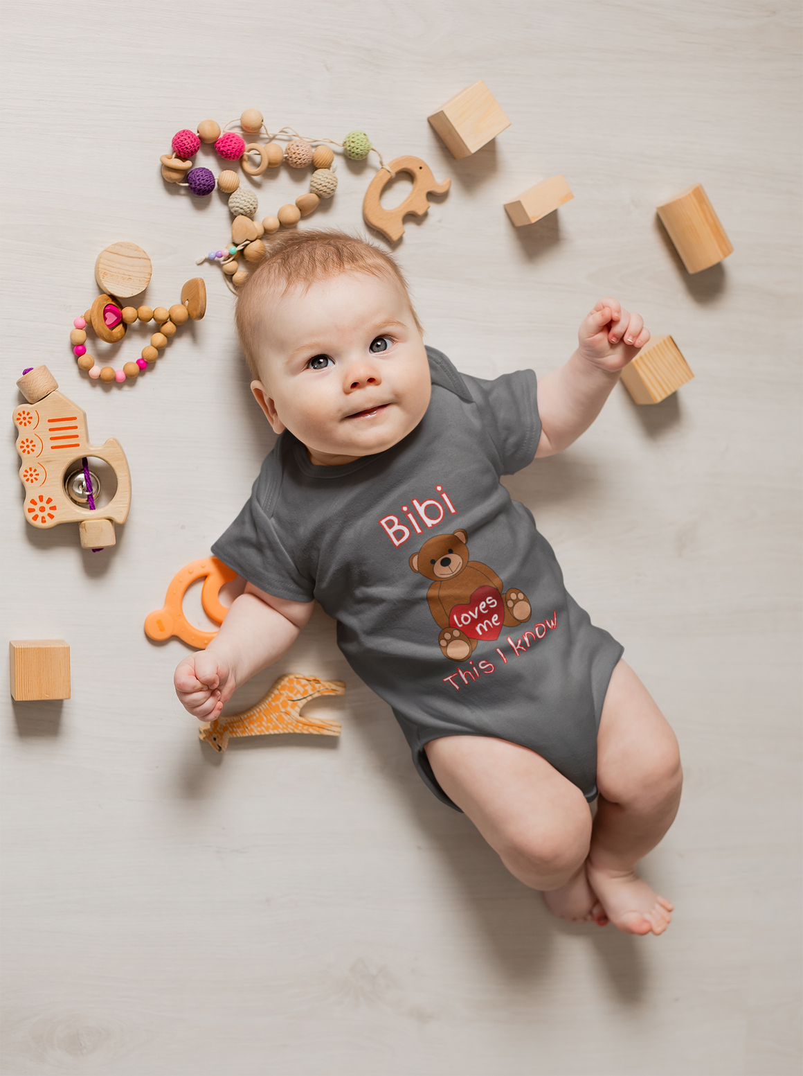 A baby lies on a wooden floor surrounded by various toys, including wooden blocks, a wooden train, and a wooden giraffe. The baby is wearing a gray onesie with a teddy bear design and the text "Bibi loves me this I know".